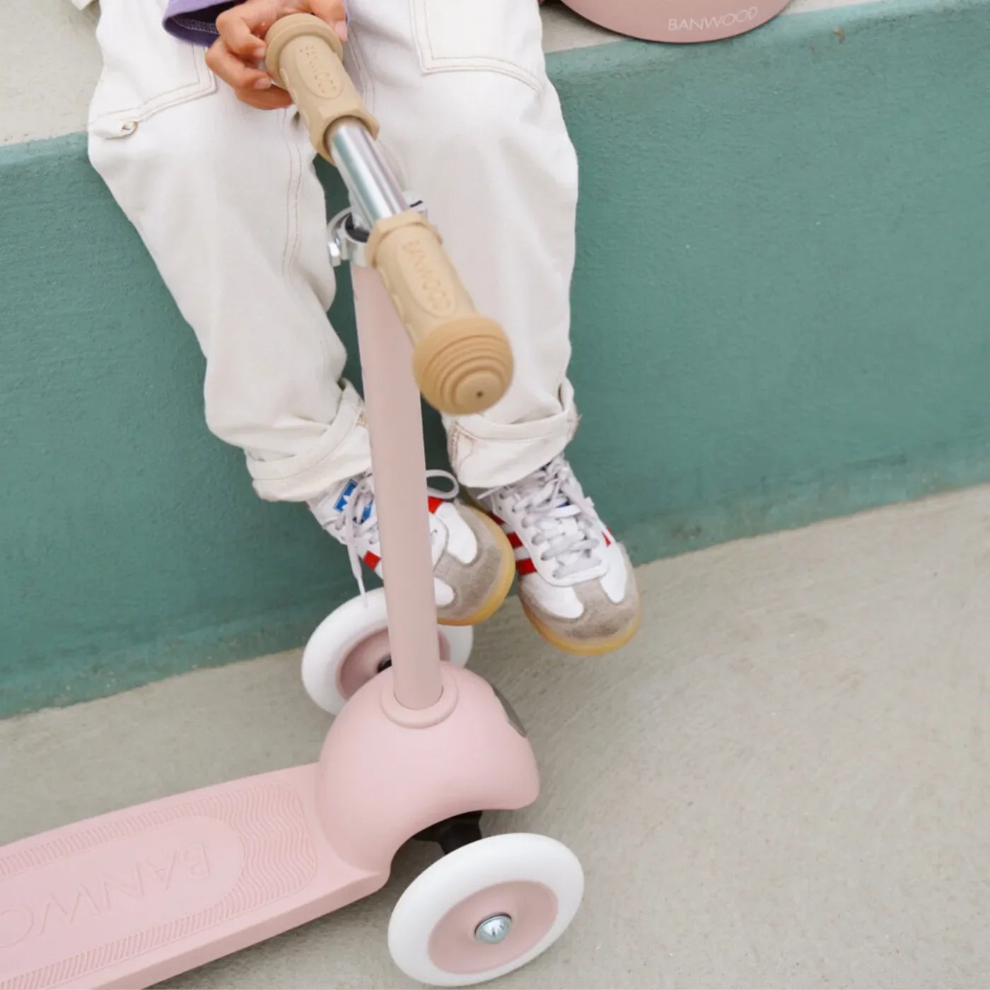 Person sitting on a pink scooter with wooden handlebar, wearing white pants and sneakers.