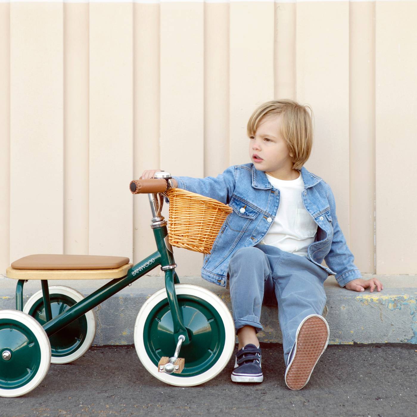 Child sitting on a tricycle with a basket against a beige wall.
