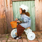 Child riding a tricycle with a basket, wearing a helmet and denim jacket, against a wooden and green curtain background.