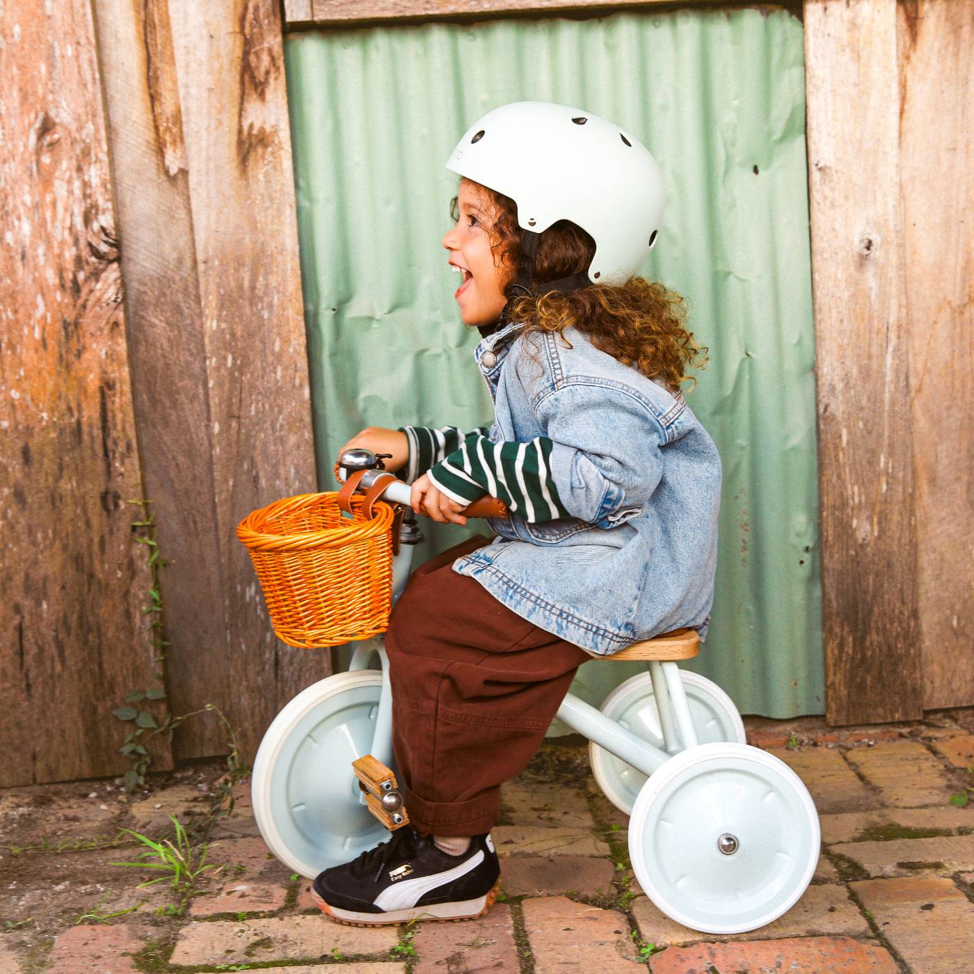 Child riding a tricycle with a basket, wearing a helmet and denim jacket, against a wooden and green curtain background.