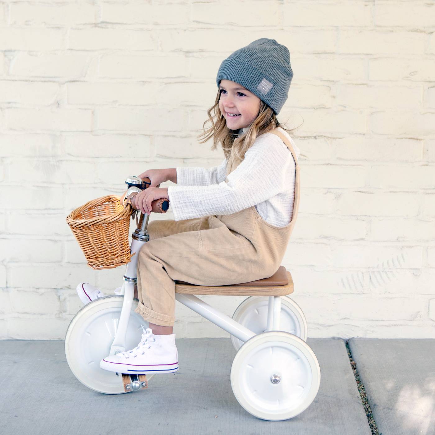 Child riding a tricycle with a basket against a light brick wall.